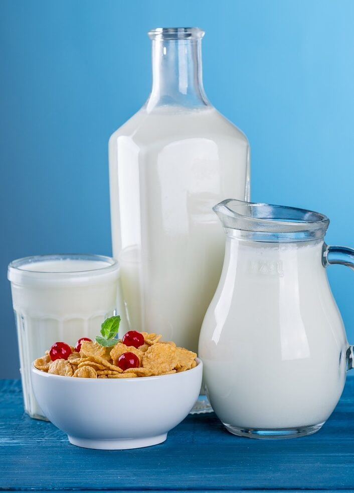 "A glass bottle, a jug, and a glass filled with fresh milk, accompanied by a bowl of cereal topped with red berries, set against a vibrant blue background. The image highlights the nutritional benefits of milk as a key part of a healthy breakfast, aligning with the theme of milk as a nutritional powerhouse for a healthy body."