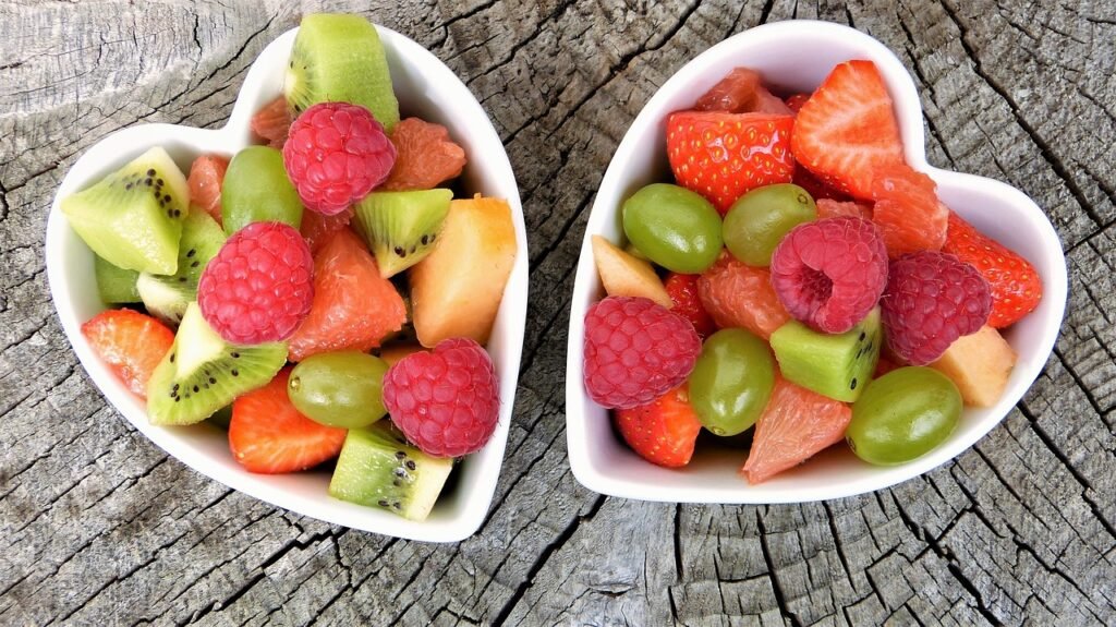 Two heart-shaped bowls filled with fresh, colorful fruits including kiwi, raspberries, strawberries, grapes, and grapefruit on a rustic wooden surface, symbolizing healthy eating for physical and mental well-being.